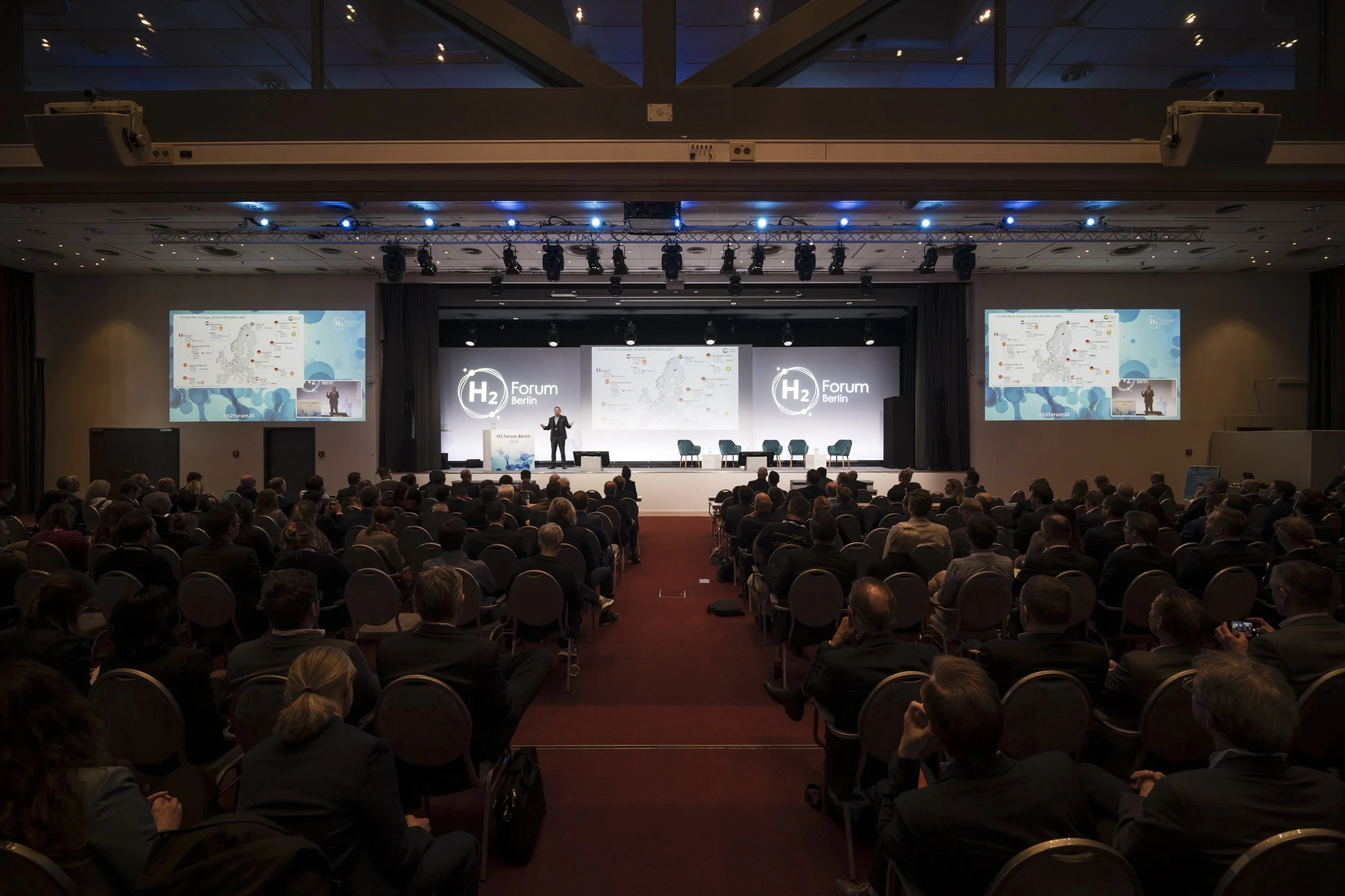 Conference hall at H2 Forum Berlin 2026: a speaker on stage addresses a seated audience, with two large screens showing a map and presentation slides.