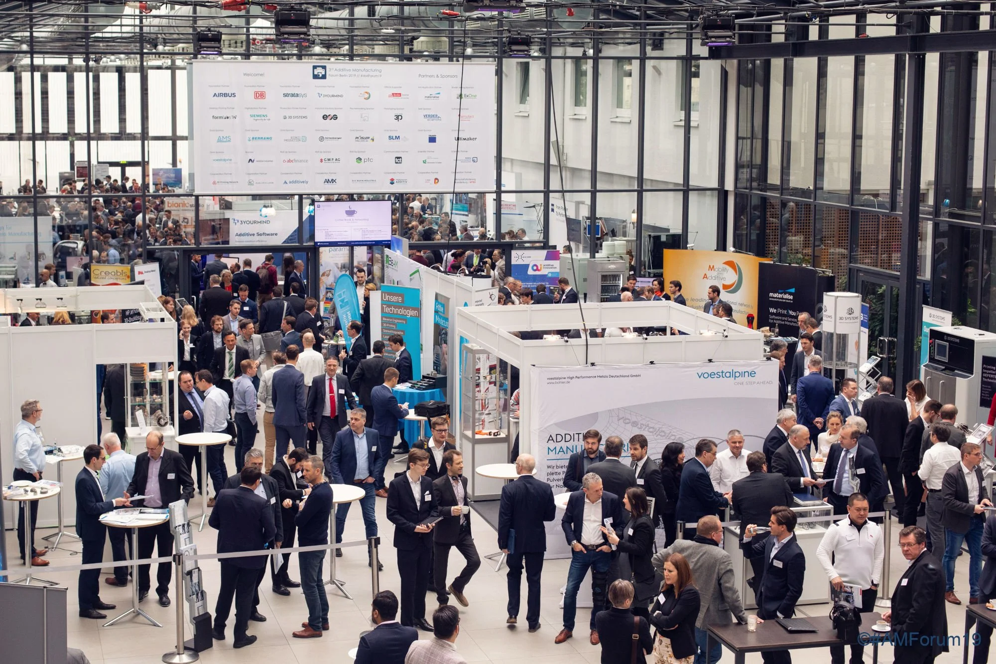 Exhibition hall at a past AM Forum, with exhibitor booths, a sponsor board and business attendees in conversation.