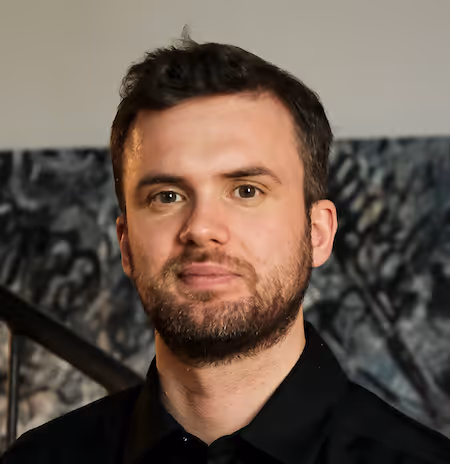 Portrait of a man with short dark hair and beard, wearing a black shirt with a blurred abstract background.