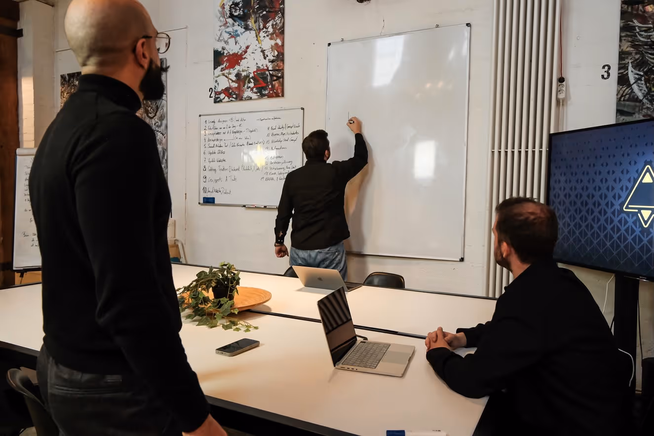 Three men in a meeting room, one writing on a whiteboard while the others watch, with laptops and a plant on the table.
