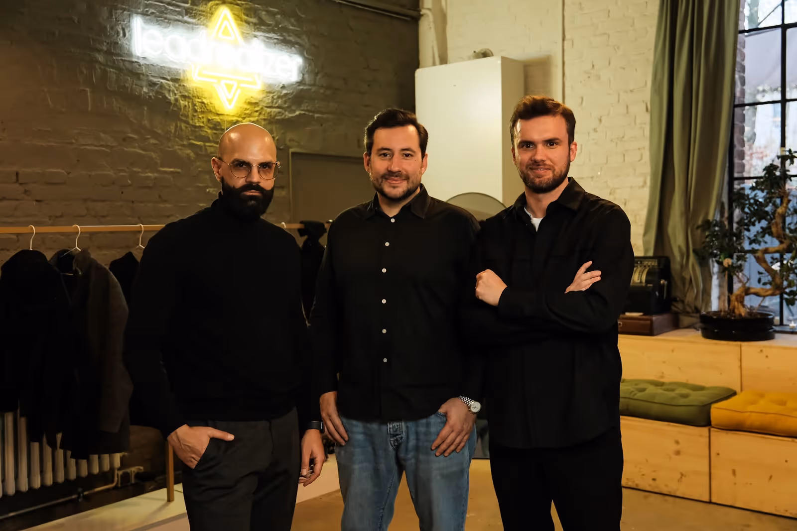 Three men standing indoors in front of a neon 'leadrealizer' sign, dressed in dark casual clothes.