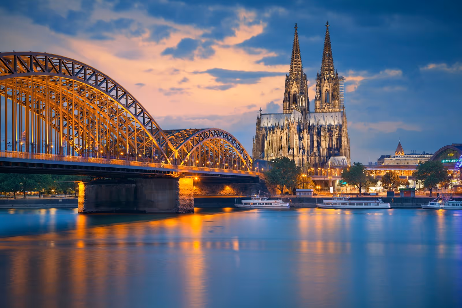 Evening view of Cologne Cathedral and the illuminated Hohenzollern Bridge over the Rhine River with boats.