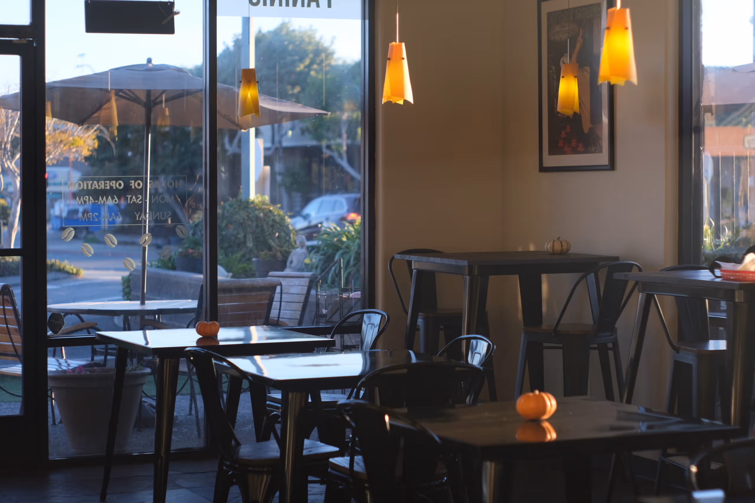 Empty coffee shop interior with small orange pumpkins on tables and large windows showing outdoor seating with umbrellas.