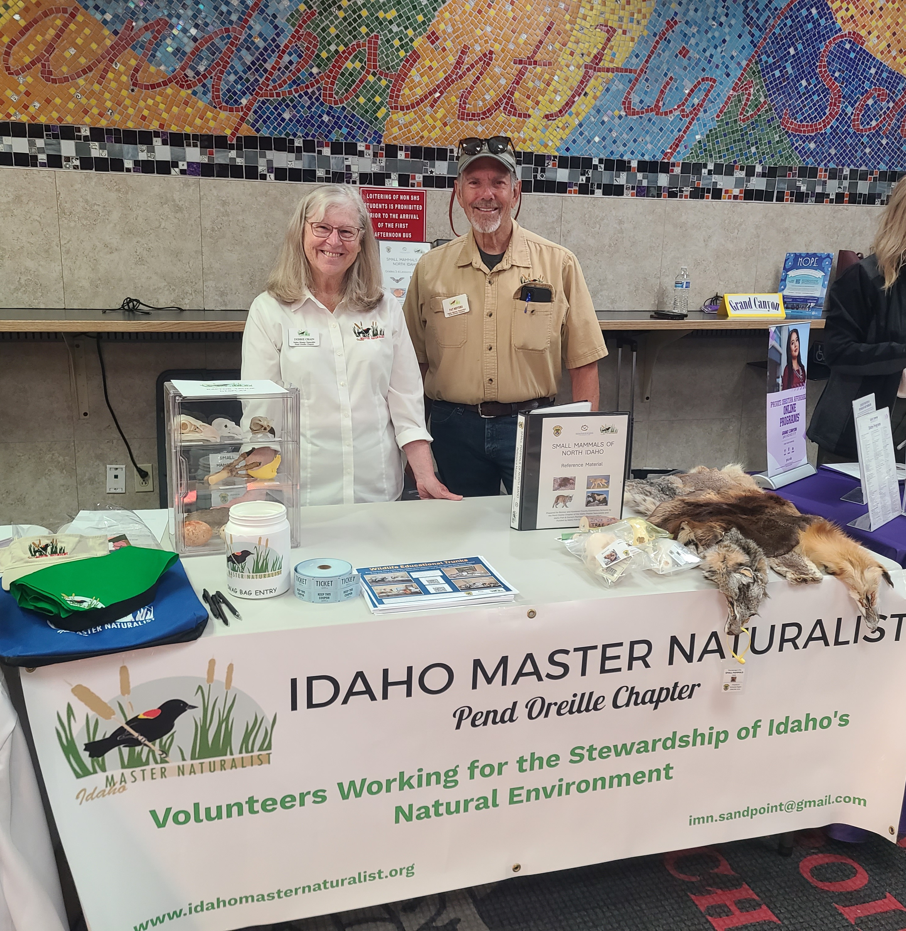 Two Idaho Master Naturalist volunteers standing behind an educational booth featuring a table of wildlife artifacts and a branded banner.