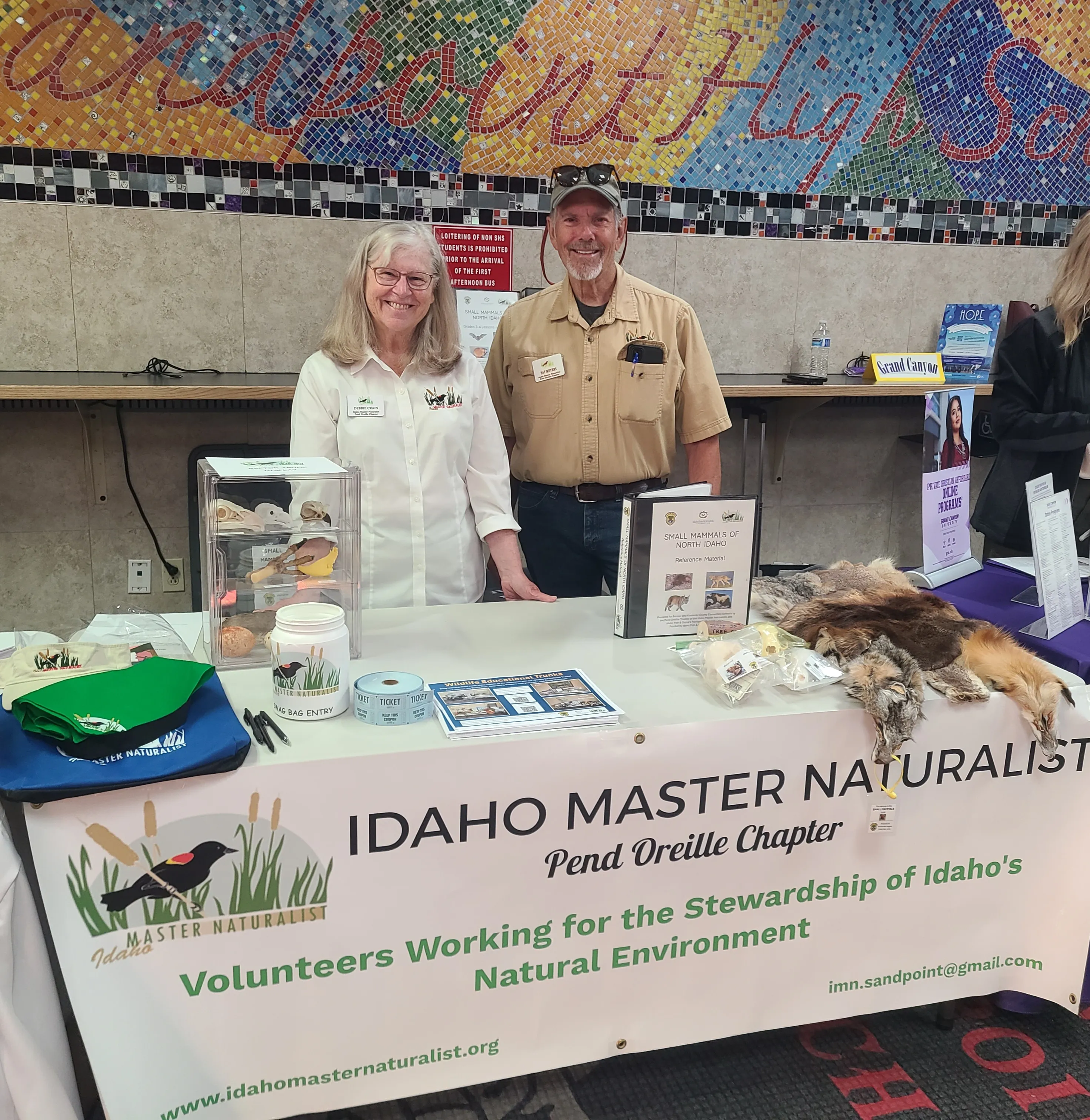 Two Idaho Master Naturalist volunteers standing behind an educational booth featuring a table of wildlife artifacts and a branded banner.