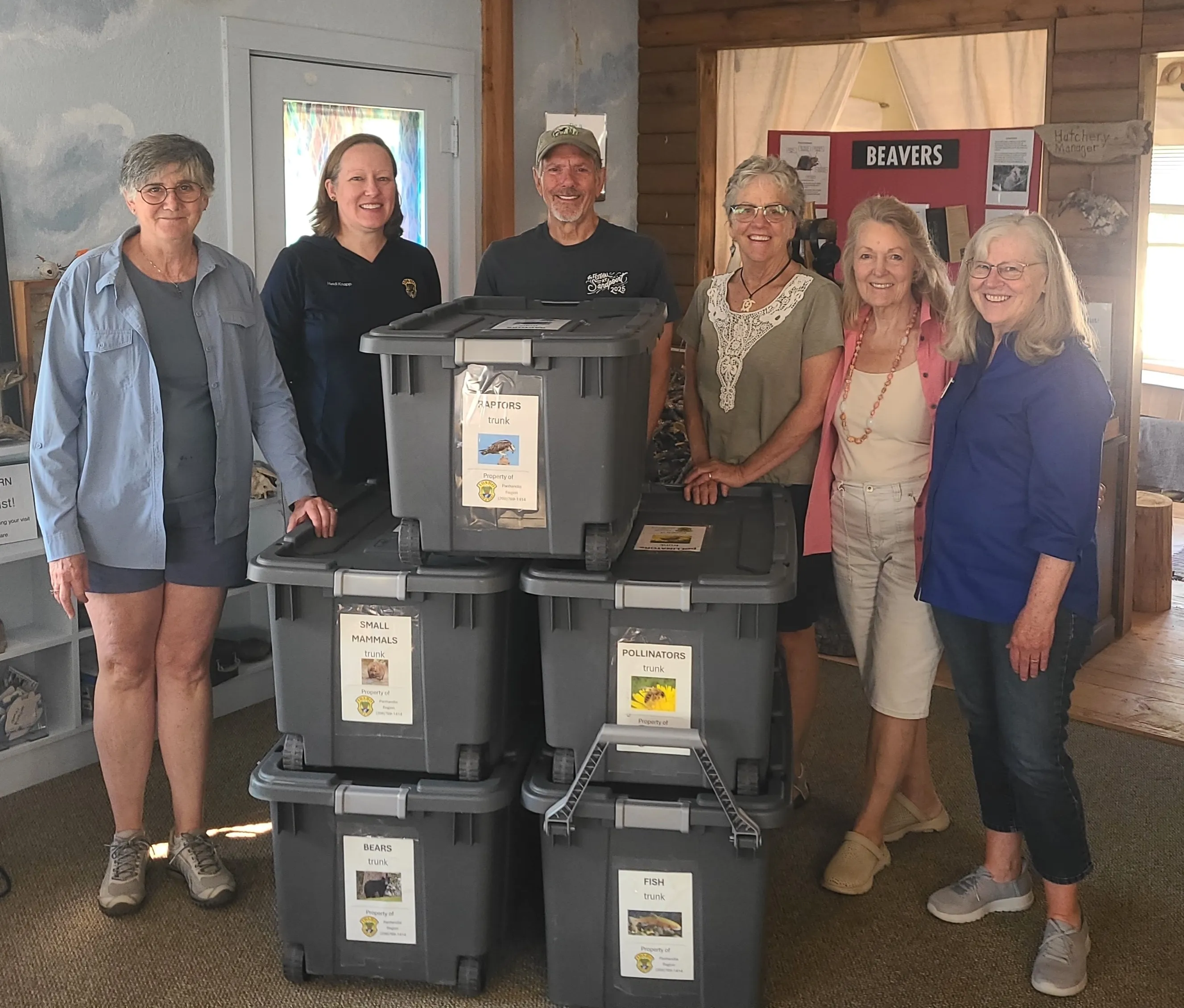 A group of volunteers posing with a stack of large, black storage bins labeled as educational wildlife trunks.