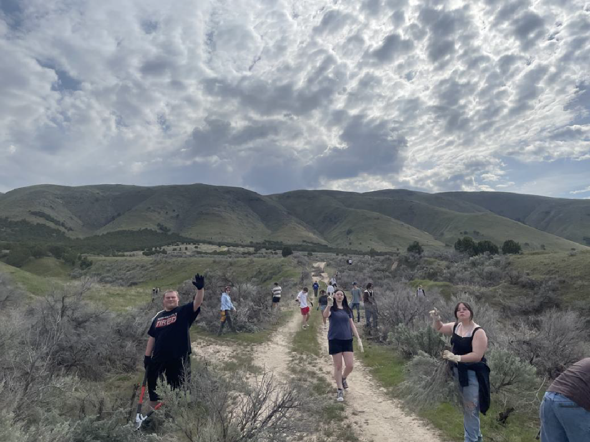 A group of people hiking along a trail through a sagebrush landscape toward rolling hills under a cloudy sky.