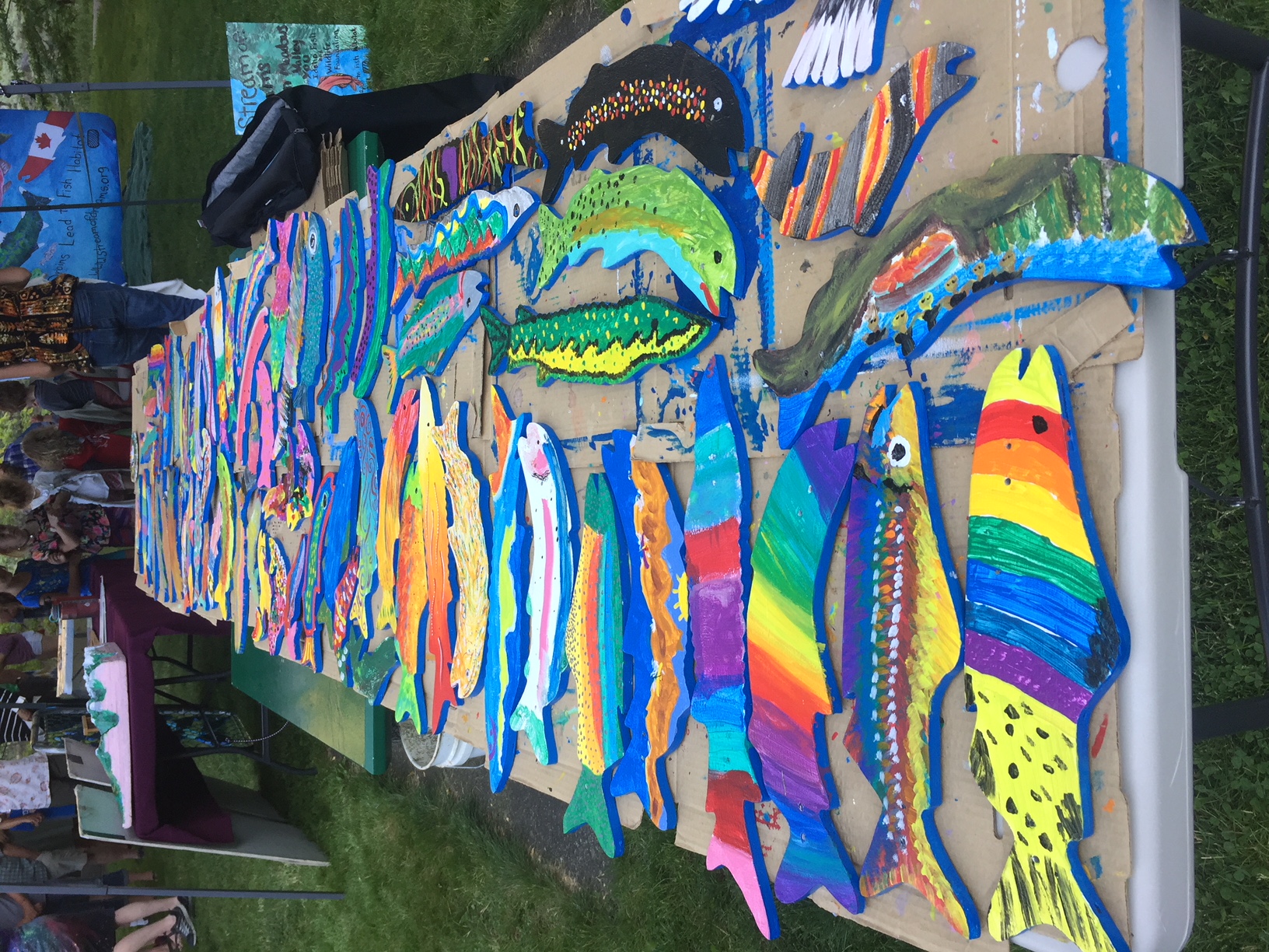 A colorful display of wooden fish painted with vibrant patterns by students, lined up on a wooden railing as part of an environmental art project.