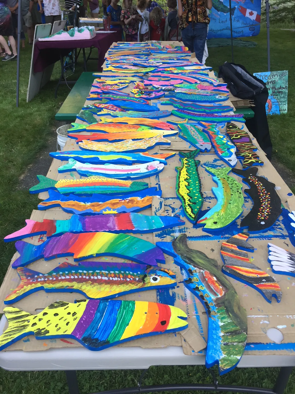 A colorful display of wooden fish painted with vibrant patterns by students, lined up on a wooden railing as part of an environmental art project.