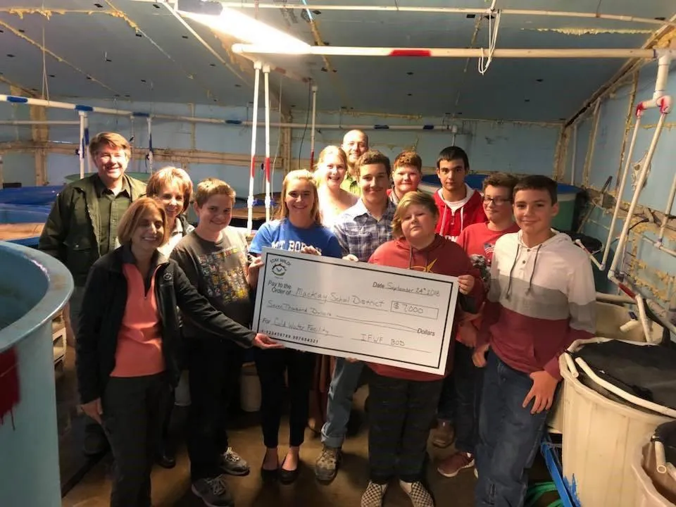 A group of students and partners standing together in a workshop holding a large ceremonial grant check from the Idaho Fish & Wildlife Foundation.