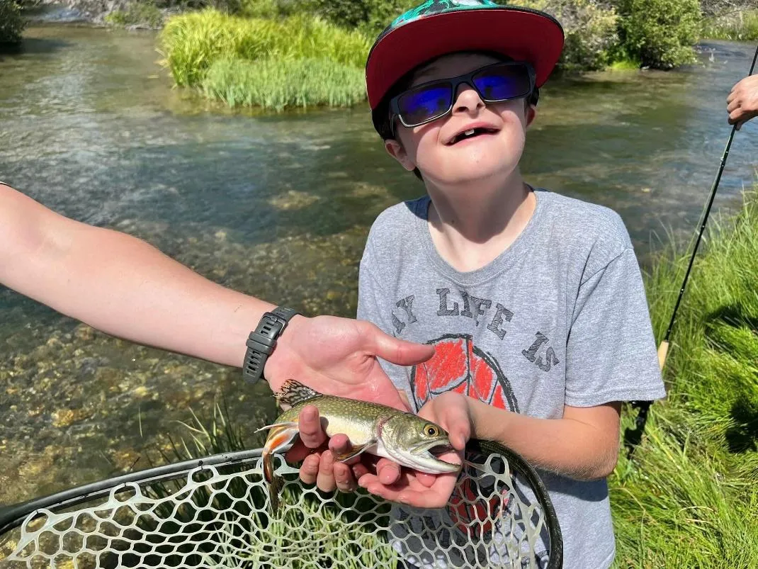 A smiling child in a hat and sunglasses holding a small trout over a fishing net on a sunny day at a riverbank.