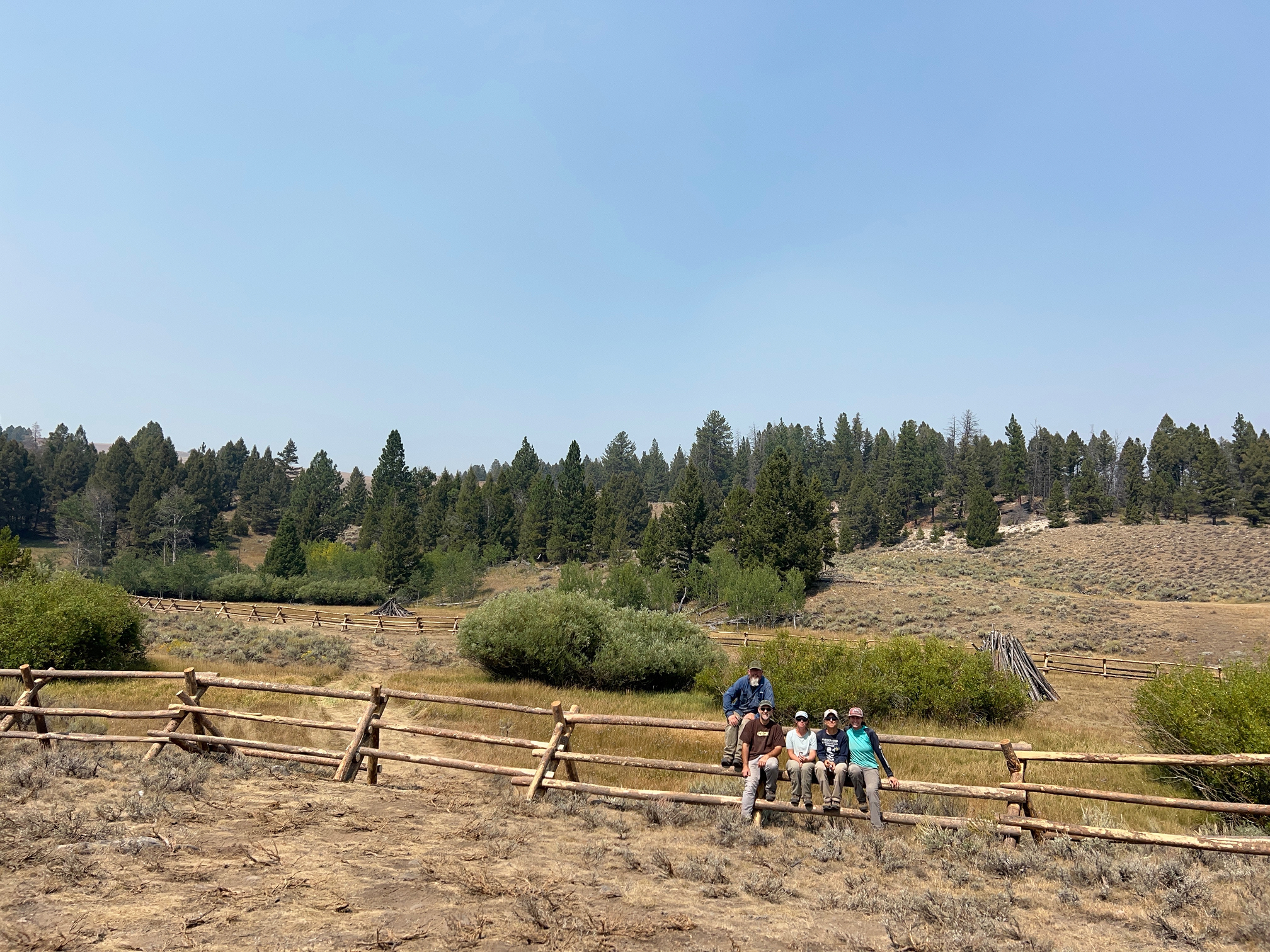 A scenic view of Moyer Basin featuring a traditional wooden buck-and-rail fence spanning an open meadow with a dense evergreen forest in the background.