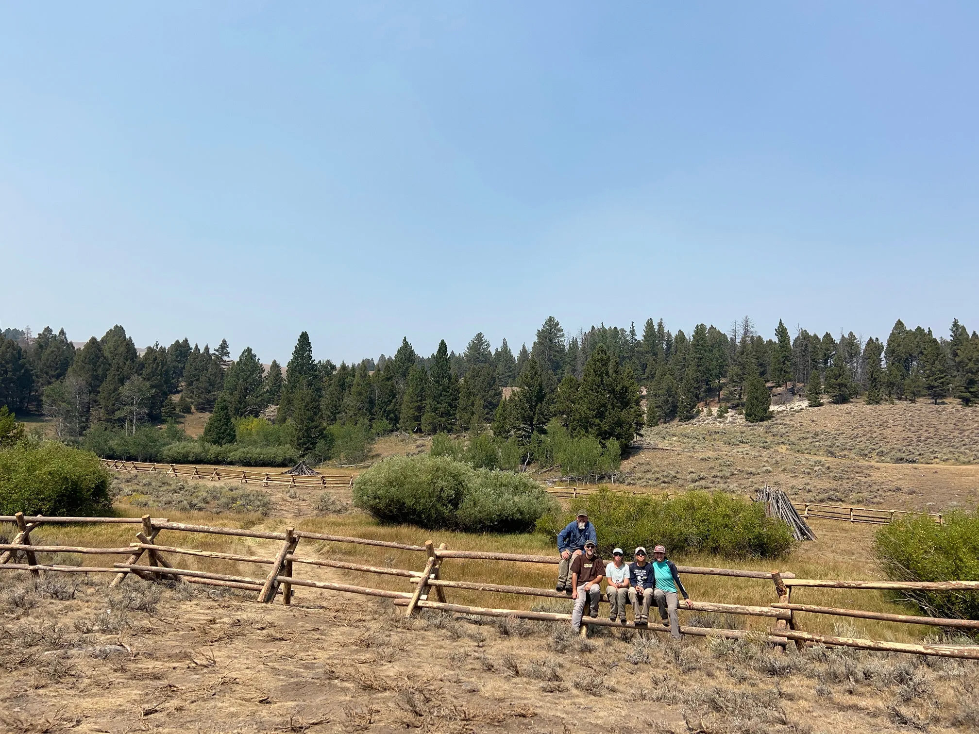 A scenic view of Moyer Basin featuring a traditional wooden buck-and-rail fence spanning an open meadow with a dense evergreen forest in the background.