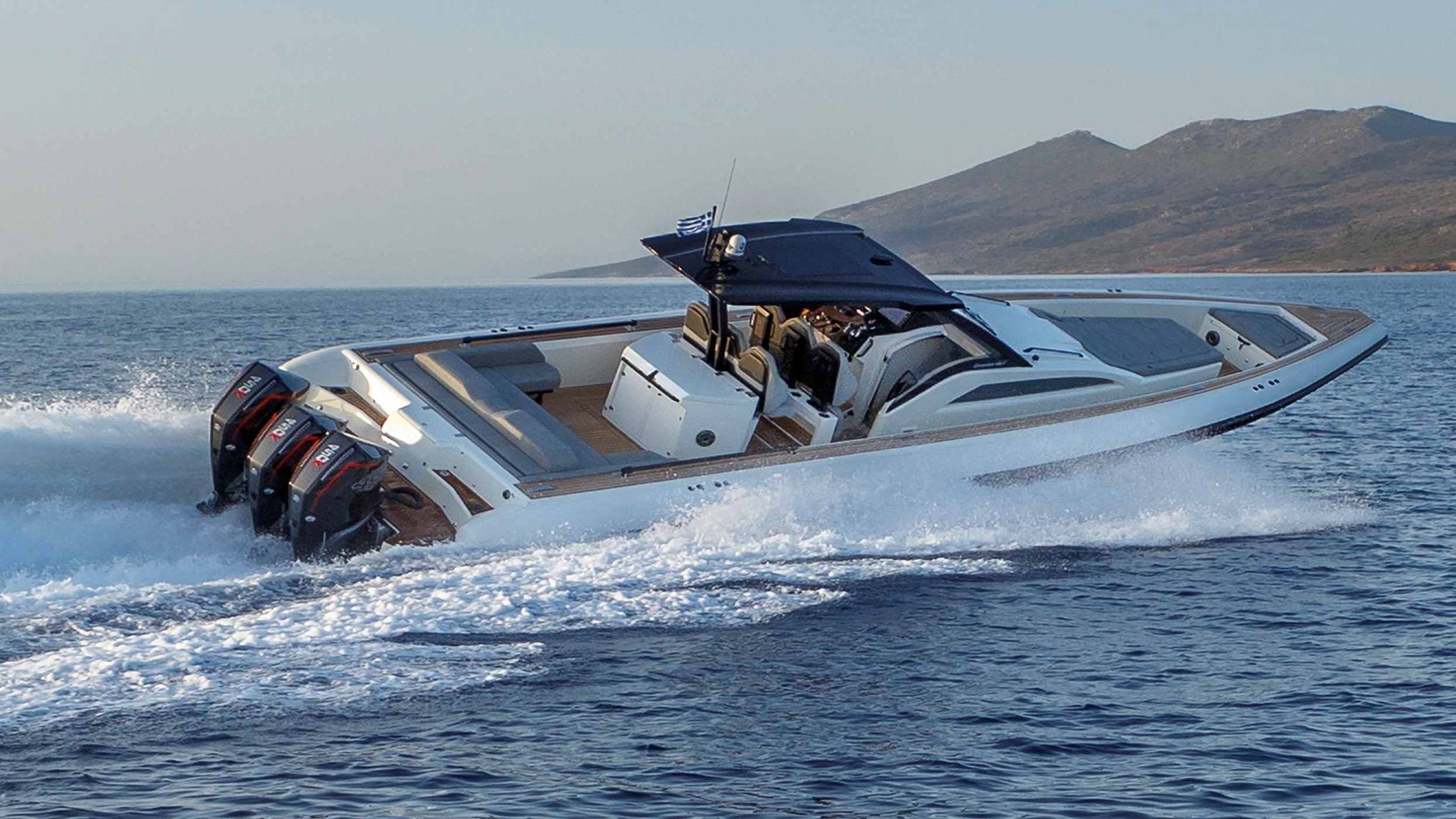 White motorboat with three outboard engines speeding on the water near a mountainous coastline.