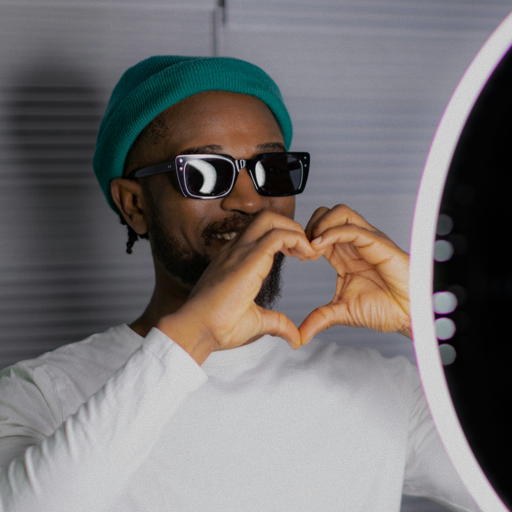 Man wearing sunglasses and a teal beanie making a heart shape with his hands in front of a ring light.