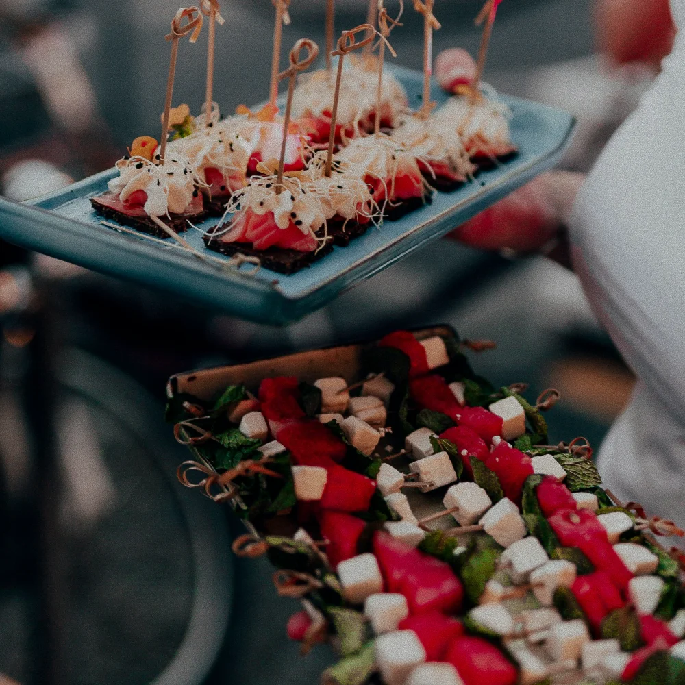 Person holding trays with assorted appetizers including skewers with cheese, watermelon, mint leaves, and crackers topped with cream and sprouts.
