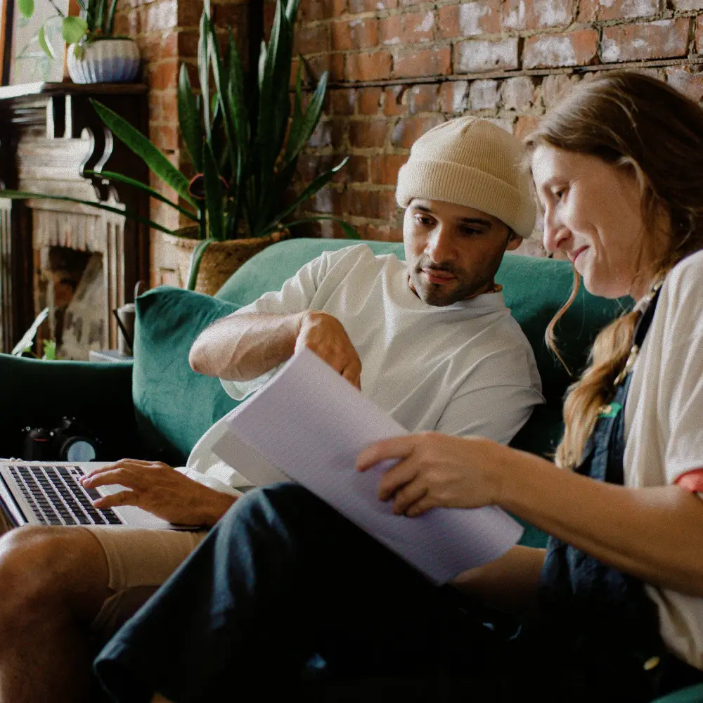 Two people sitting on a green couch reviewing notes and using a laptop in a cozy room with exposed brick walls and plants.