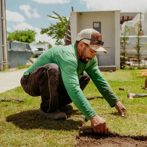 Man wearing a green long-sleeve shirt and cap laying sod on a lawn outdoors.