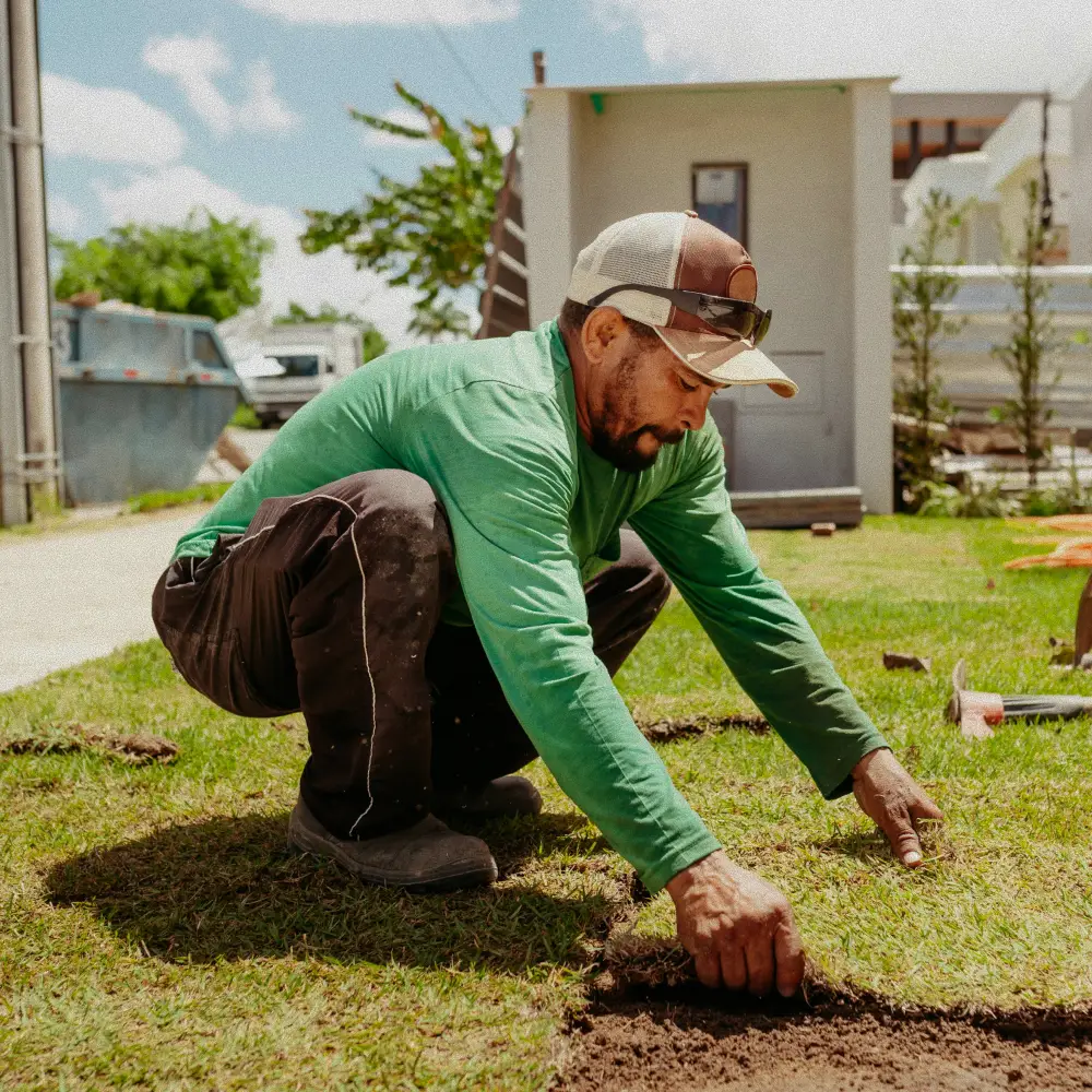 Man wearing a green long-sleeve shirt and cap laying sod on a lawn outdoors.