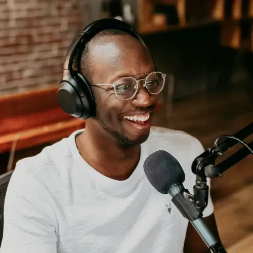 Smiling man with glasses and headphones speaking into a microphone in a recording setting.