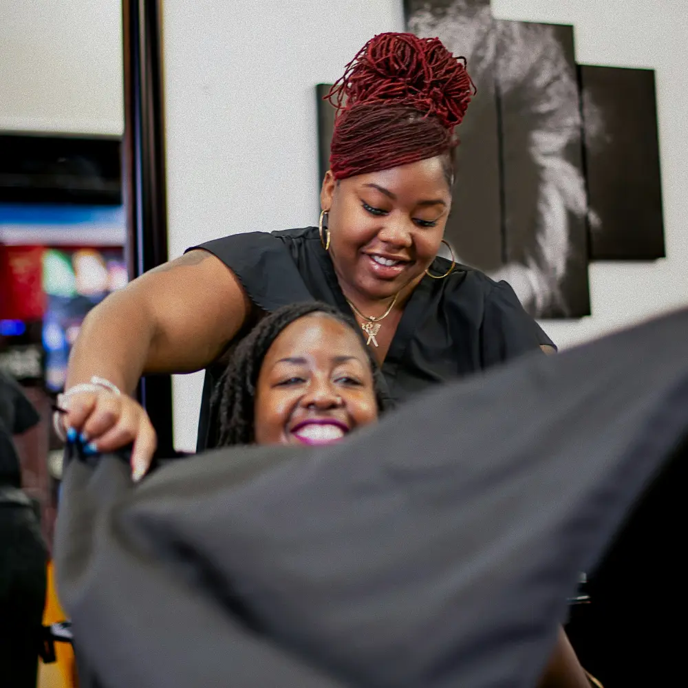 Hairdresser with red braided hair smiling while draping a black cape over a happy seated client in a salon.