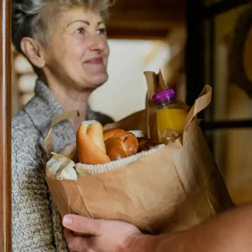Smiling elderly woman receiving a brown paper grocery bag with bread and a bottle of orange juice.