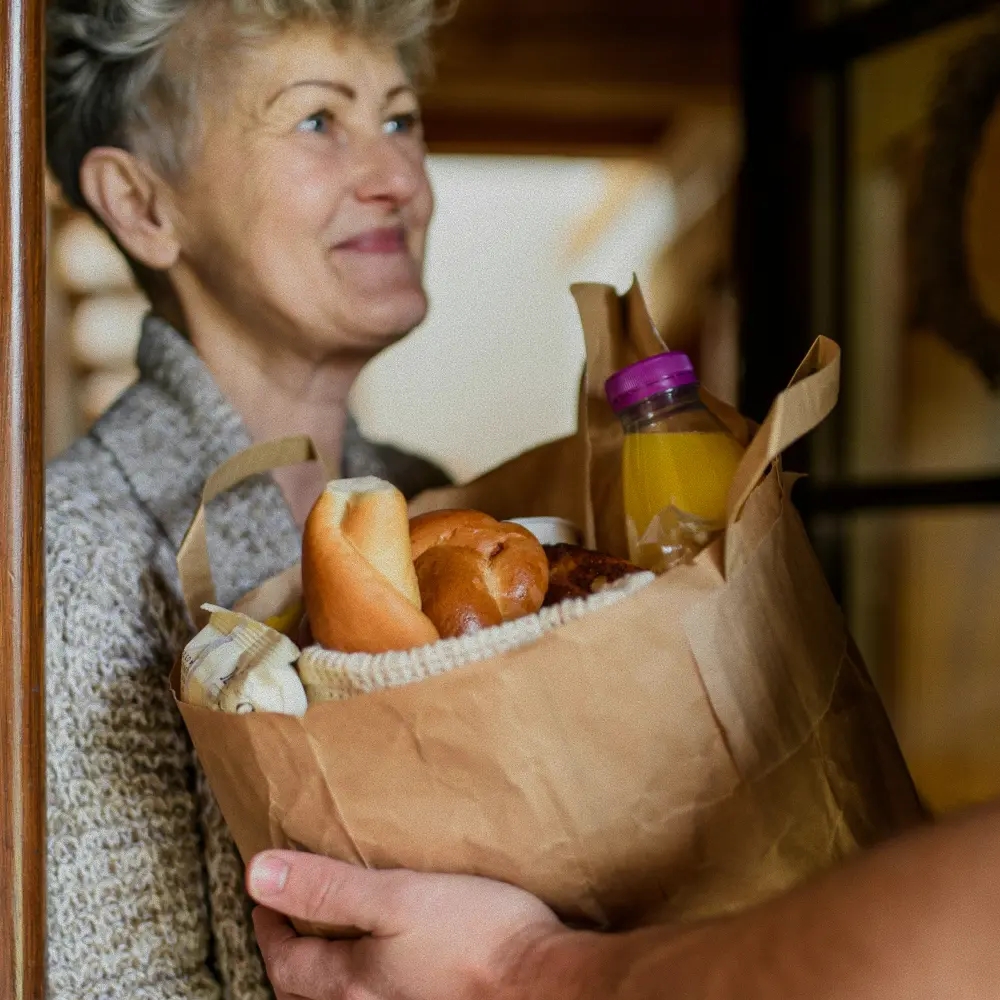 Smiling elderly woman receiving a brown paper grocery bag with bread and a bottle of orange juice.