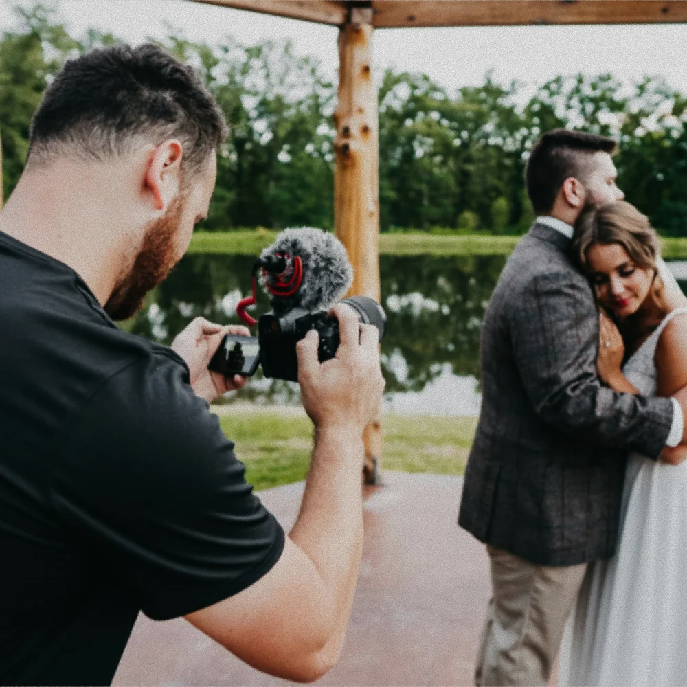 Photographer filming a couple embracing near a lake under a wooden gazebo.