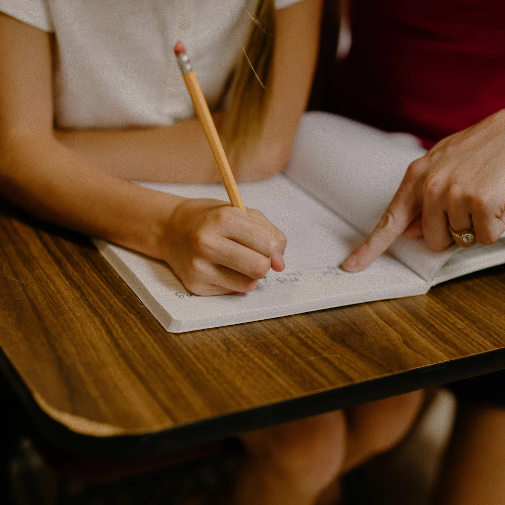 Child holding pencil writing in a notebook with an adult pointing at the writing.