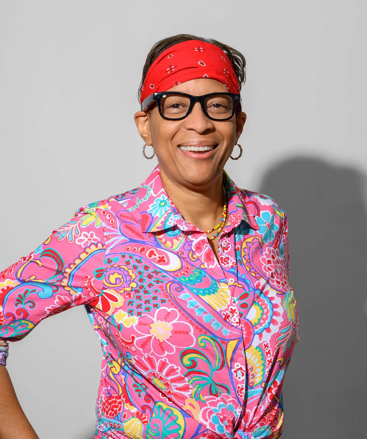 Smiling person wearing a red bandana, black glasses, hoop earrings, and a bright floral patterned pink shirt.