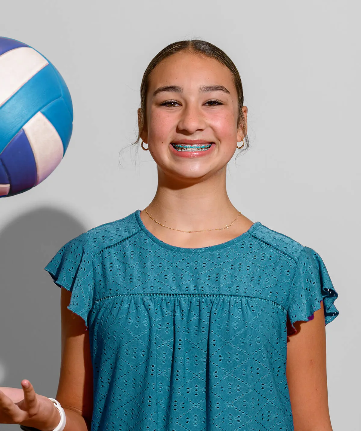 Smiling teenage girl with braces wearing a blue patterned top, holding a blue and white volleyball.