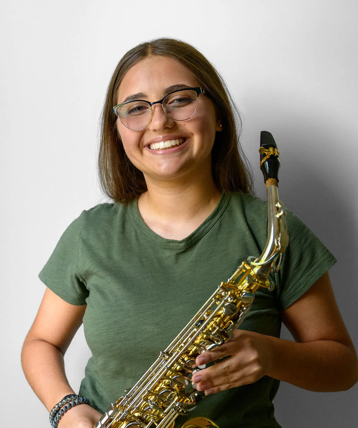 Smiling young woman wearing glasses and a green shirt holding a saxophone against a white background.