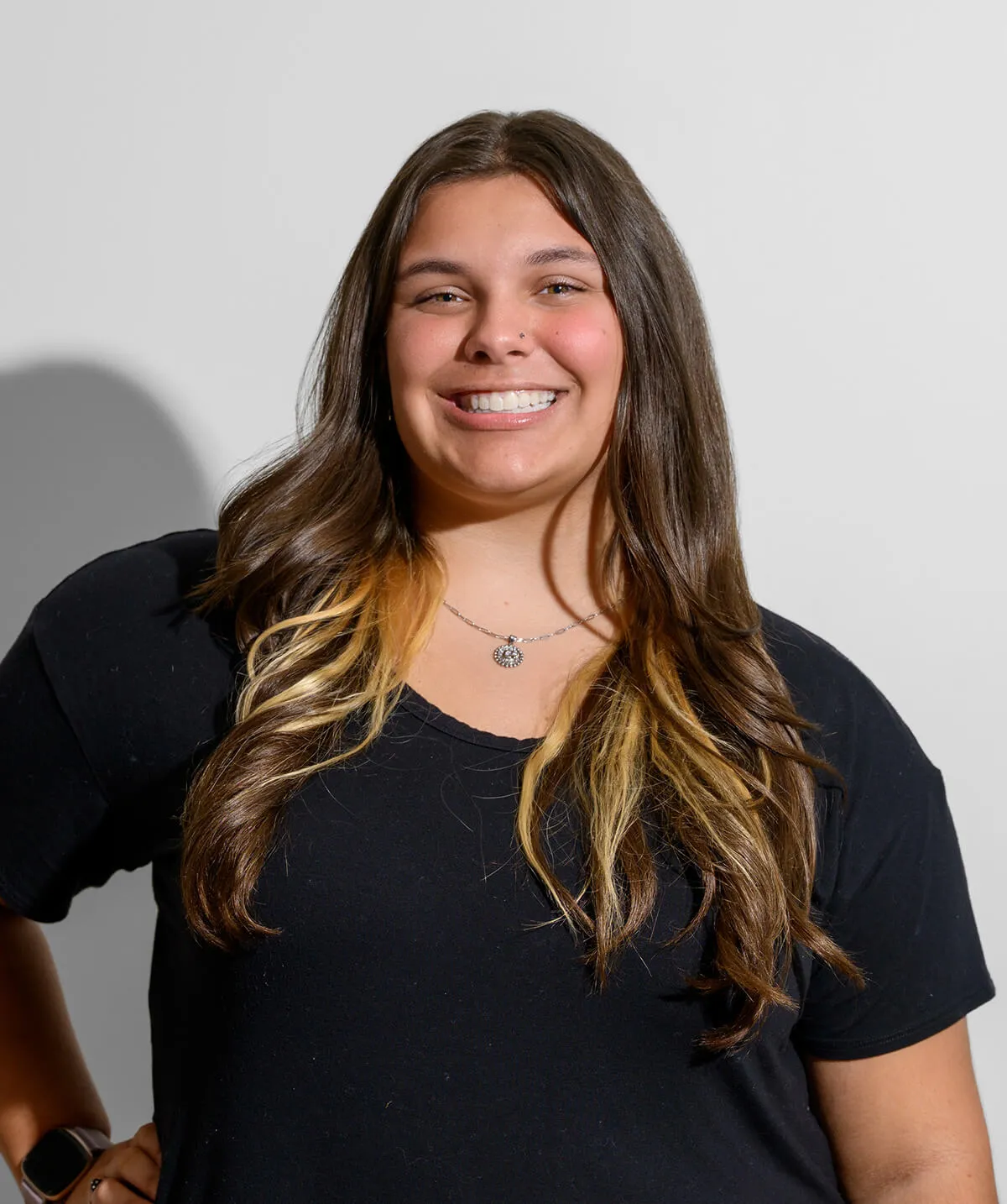 Smiling young woman with long brown hair and blonde highlights wearing a black shirt and a necklace.
