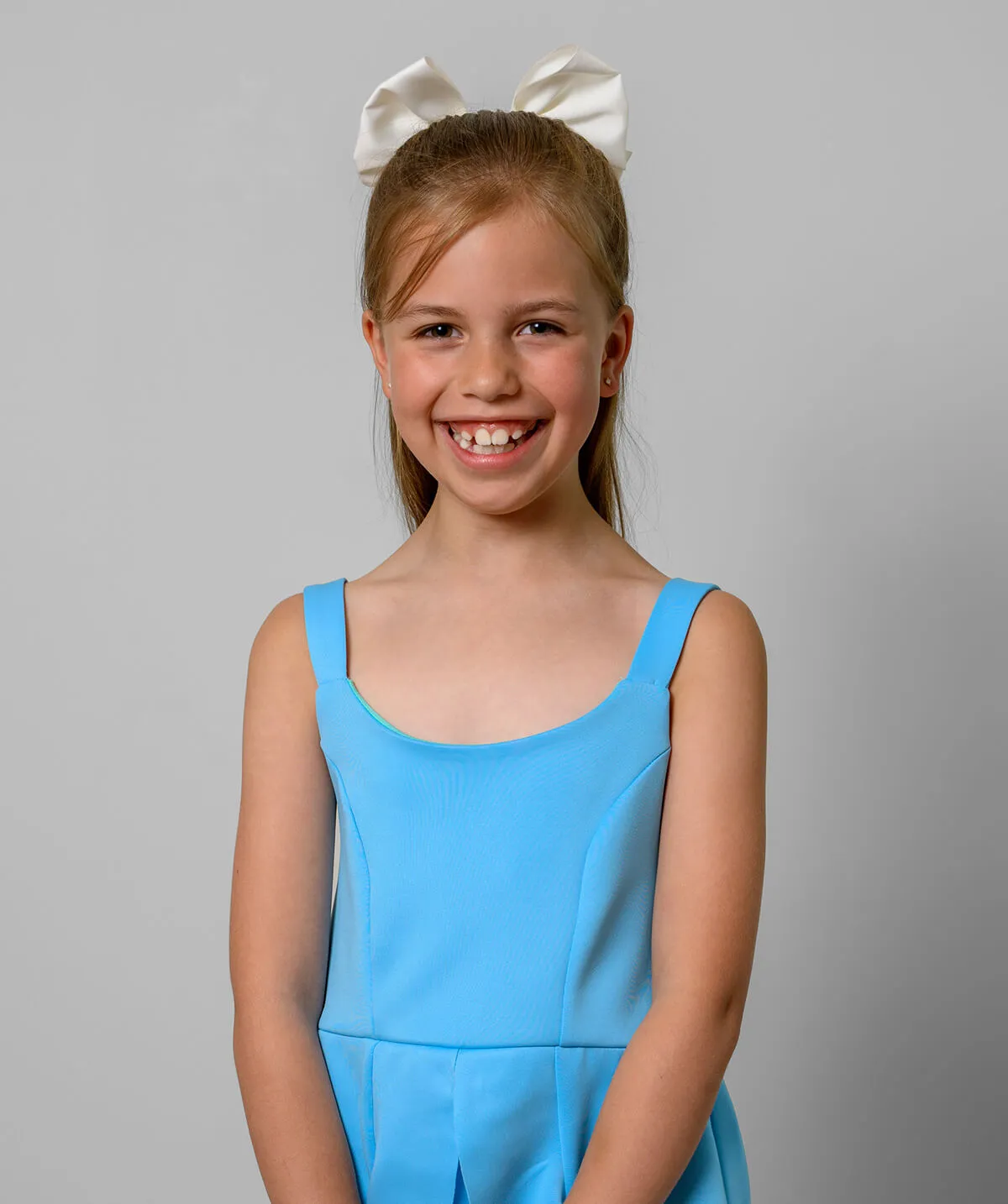 Smiling young girl with a white bow in her hair wearing a sleeveless light blue dress against a gray background.