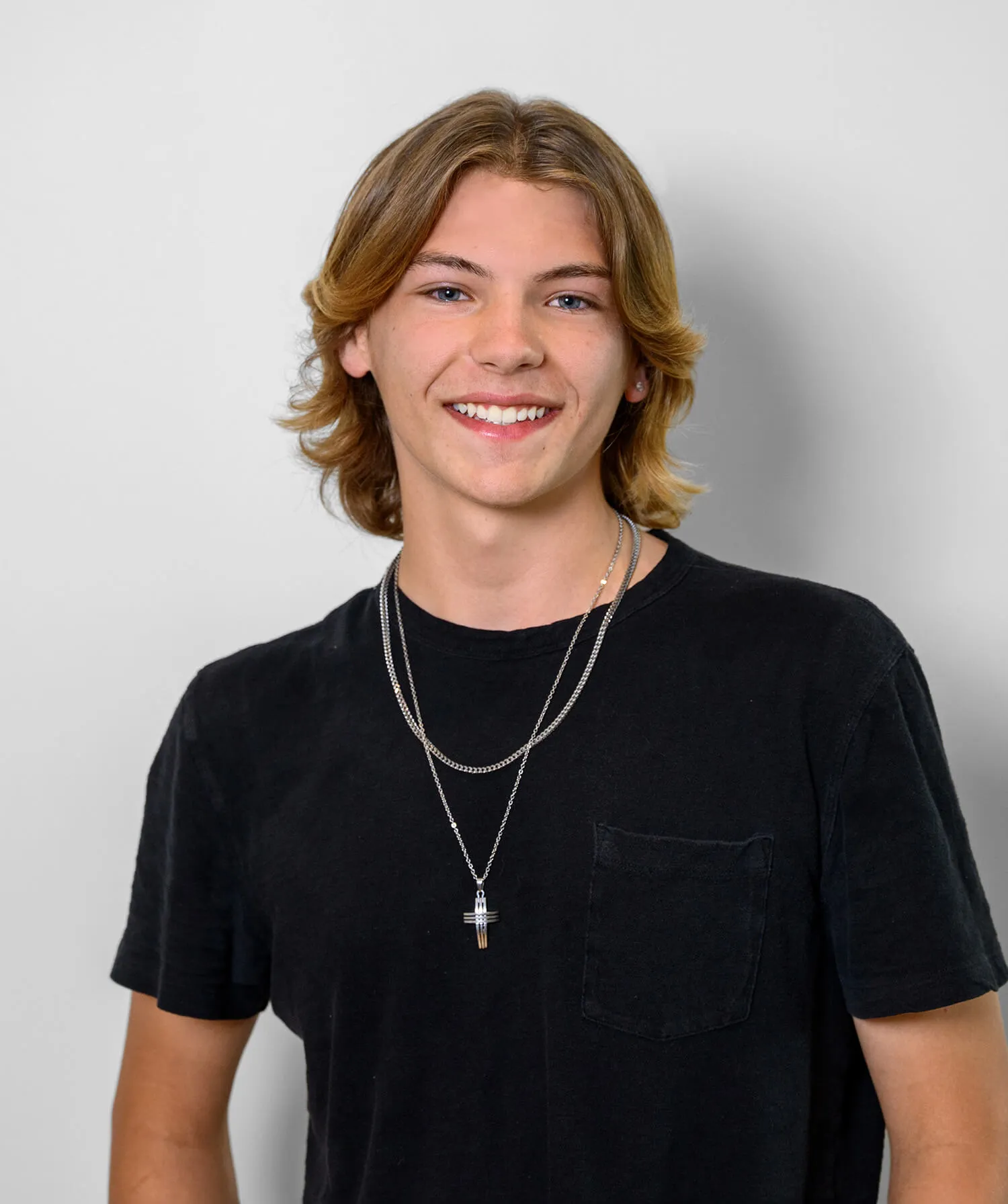 Young man with medium-length blond hair wearing a black t-shirt and silver necklaces smiling at the camera.