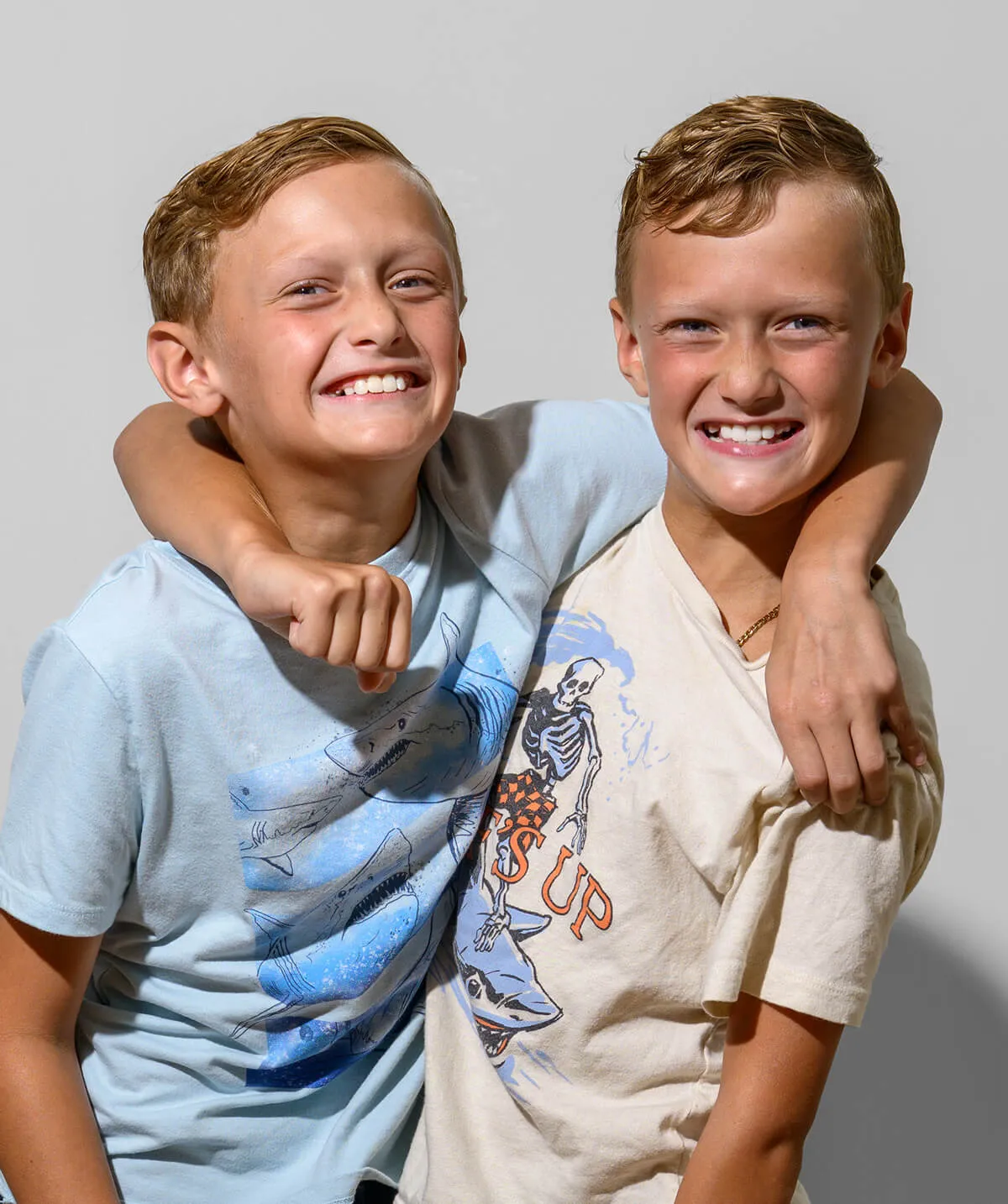Two smiling boys with light brown hair, one wearing a light blue shark graphic t-shirt and the other a beige t-shirt with a skeleton and shark design, hugging each other.