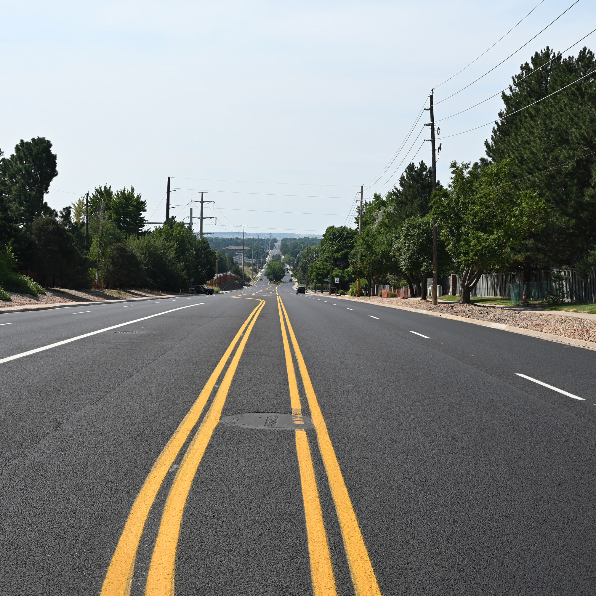 Wide multi-lane asphalt road with fresh yellow double center lines and green trees lining both sides under a clear sky.