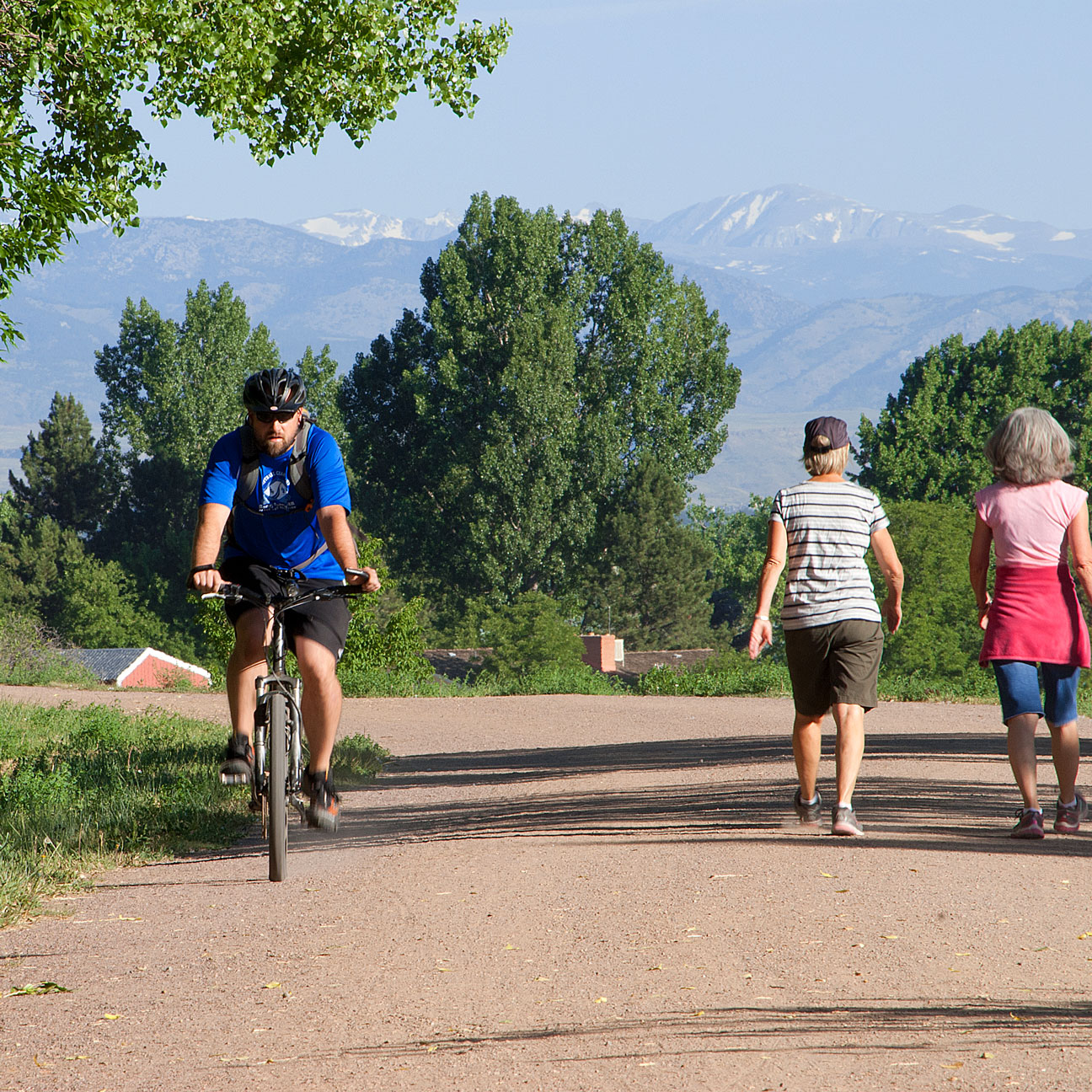 Man cycling on a gravel trail with two women walking in opposite direction, with trees and snow-capped mountains in the background.