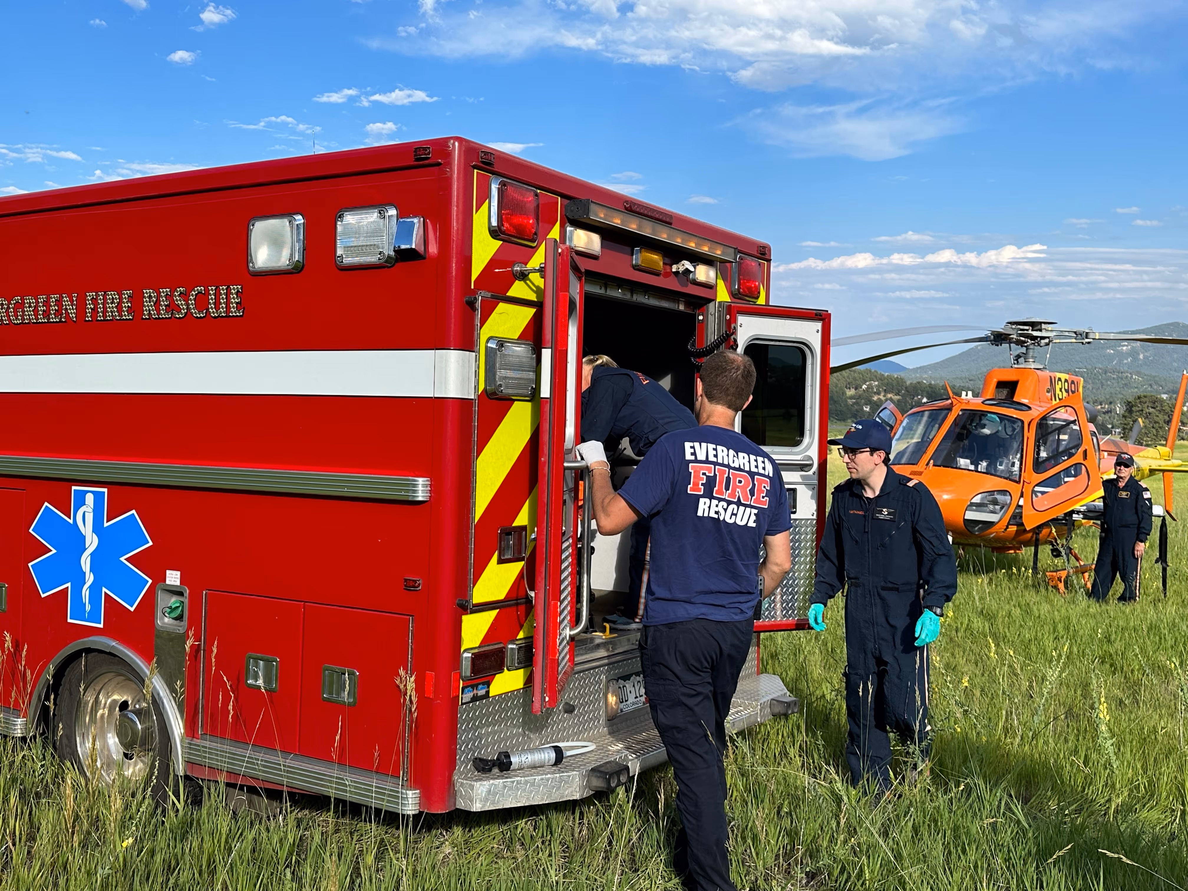 Fire rescue team members preparing equipment at the back of a red ambulance with an orange helicopter in a grassy field.