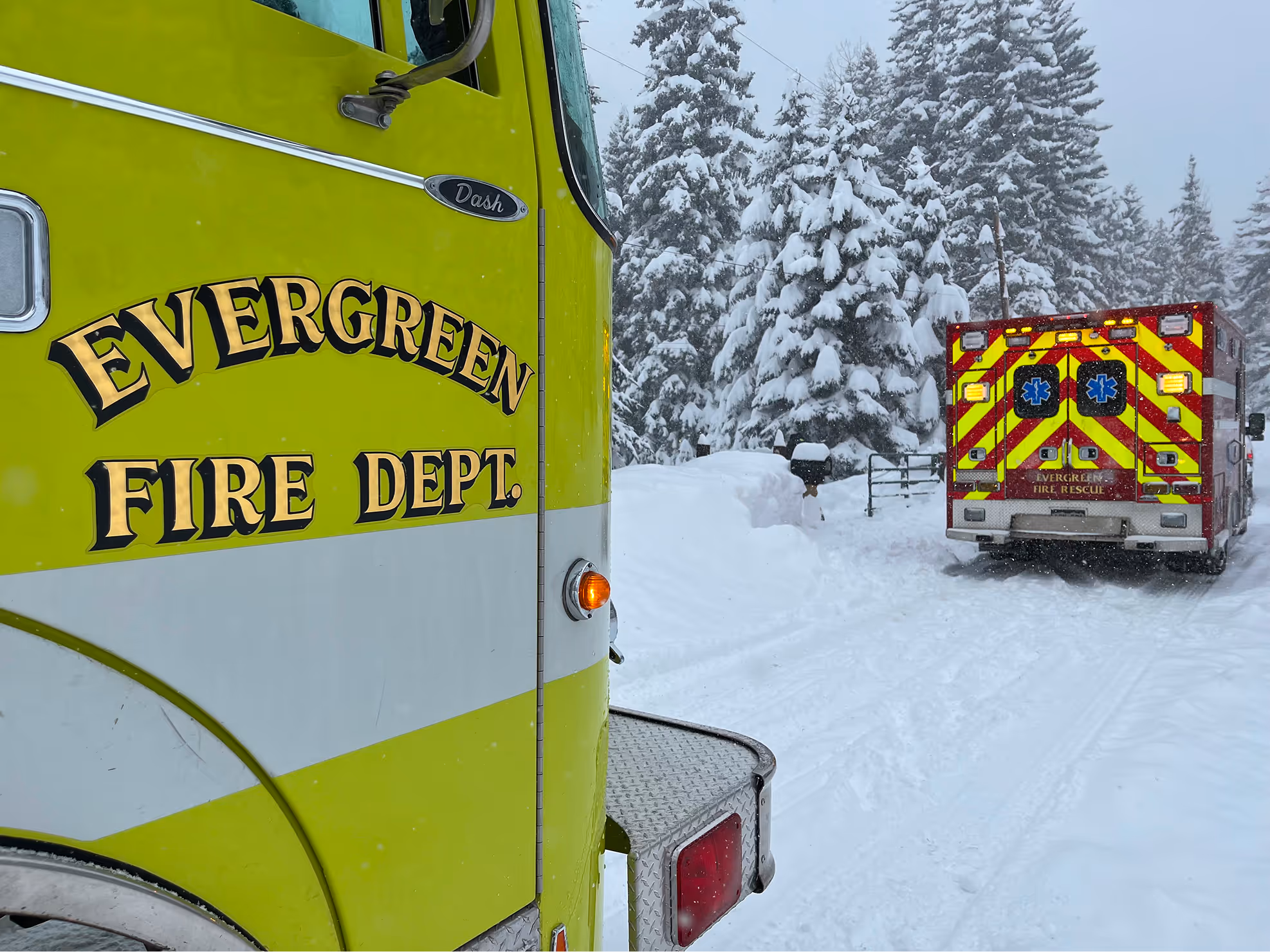 Green and white Evergreen Fire Department truck near a red and yellow-striped Evergreen Fire Rescue ambulance on a snow-covered road with snow-laden evergreen trees.