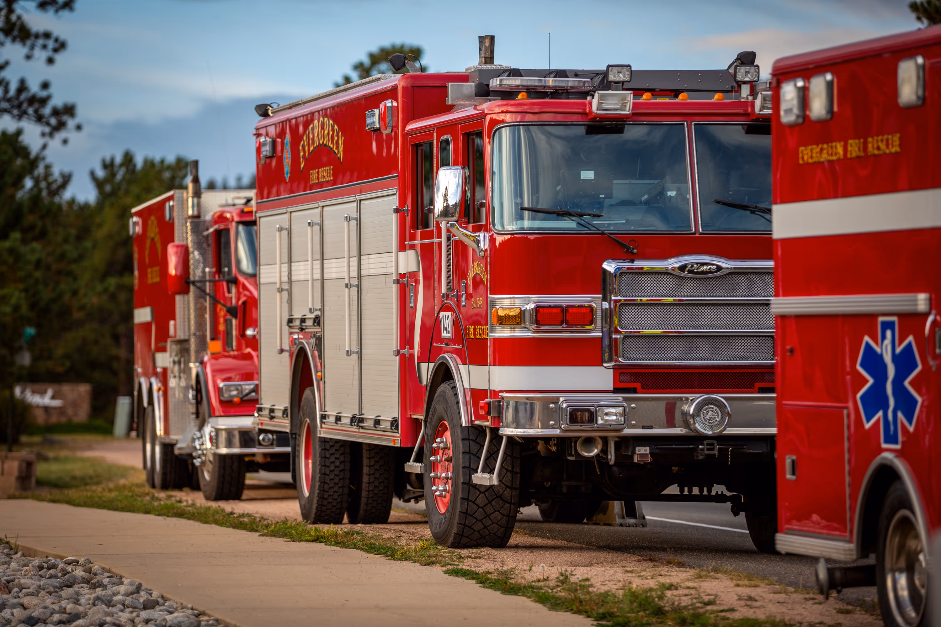 Parked red Evergreen Fire Rescue trucks and ambulance lined up on a road beside a sidewalk and trees.