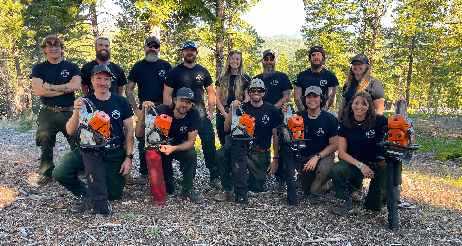 Group of eleven people in black shirts and green pants posing outdoors in a forest, some holding orange chainsaws.
