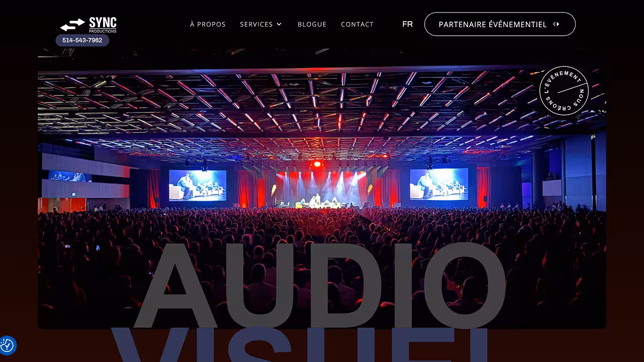 Large audience seated in a dark auditorium watching a stage with bright lights and two large screens on either side.