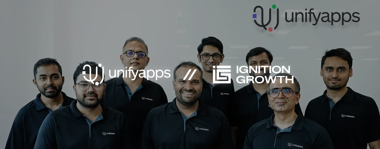 Group of eight men wearing black unifyapps shirts standing and smiling in front of a wall with unifyapps and Ignition Growth logos.
