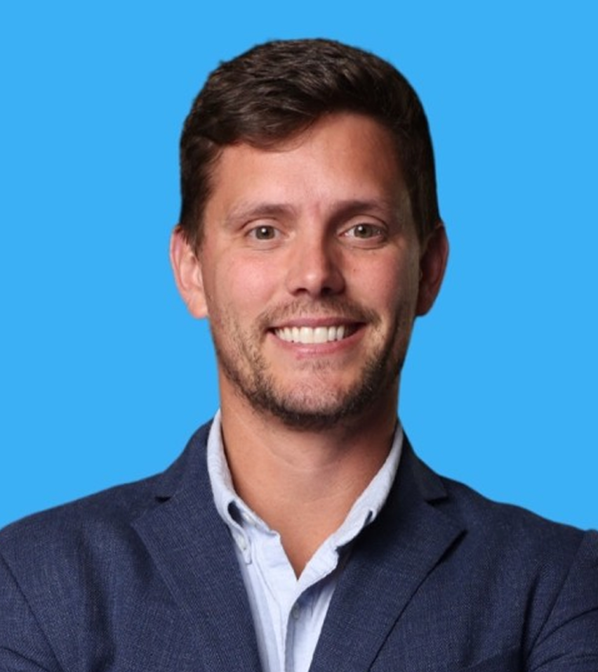 Smiling man with short brown hair wearing a blue blazer and light blue shirt against a bright blue background.