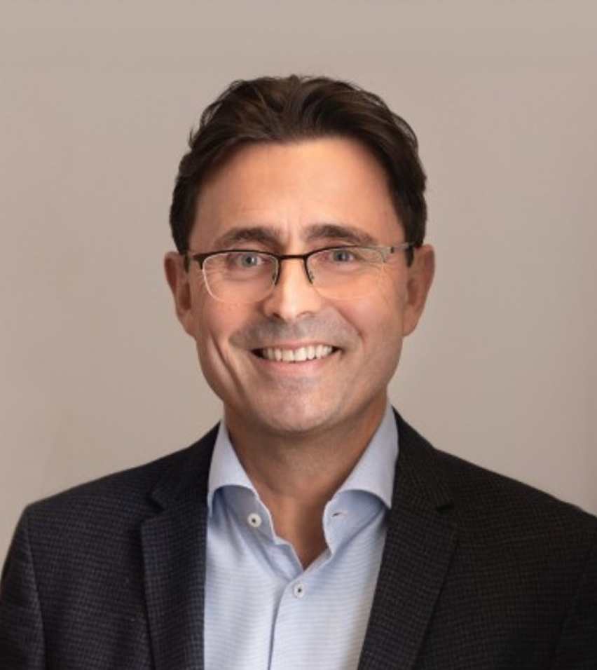 Smiling man with glasses wearing a dark blazer and light blue shirt against a plain background.