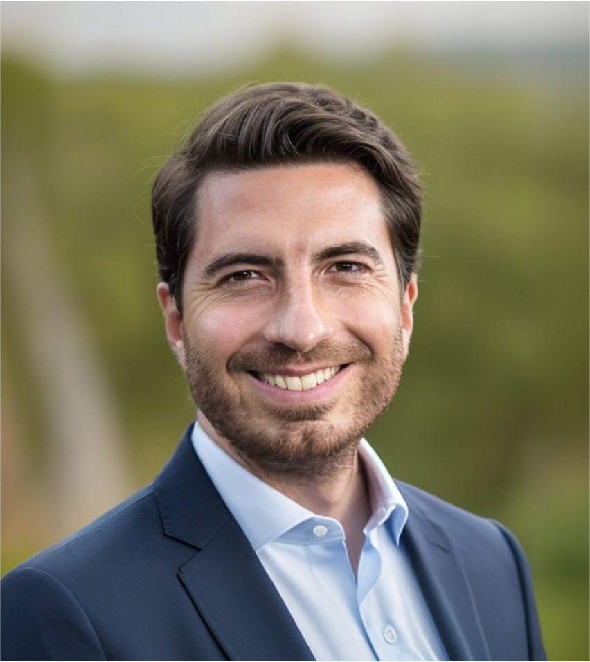 Smiling man with dark hair and beard wearing a blue suit jacket and light blue shirt, outdoors with green blurred background.