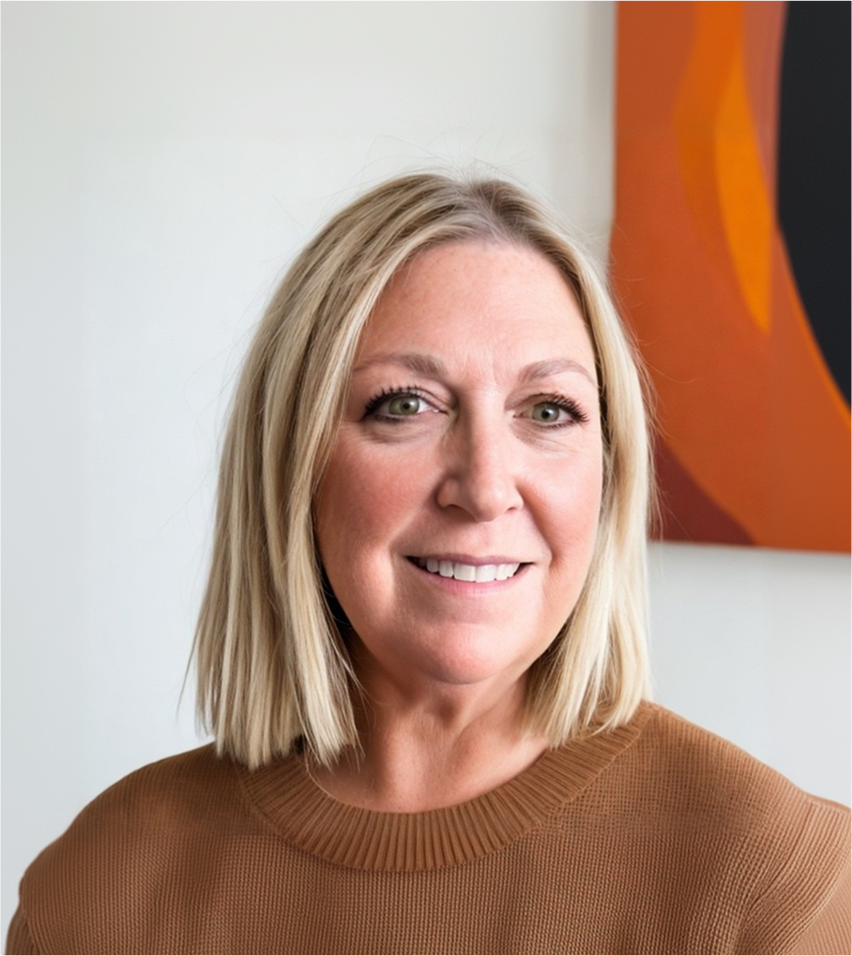Smiling blonde woman with shoulder-length hair wearing a brown sweater, standing indoors against a white wall with abstract orange and black artwork.
