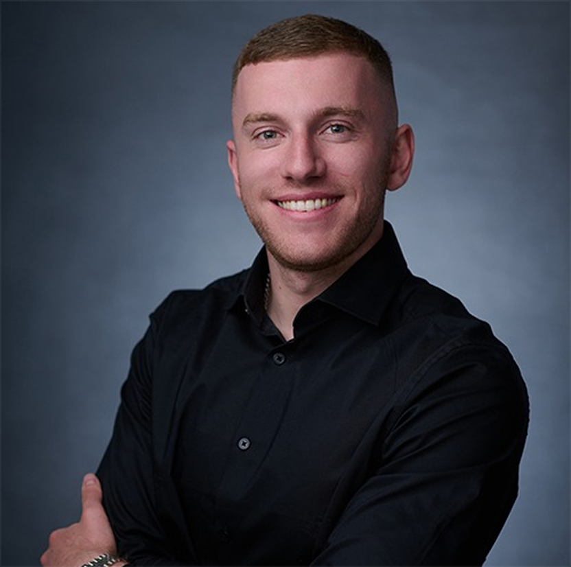 Smiling young man with short hair wearing a black button-up shirt against a gray background.