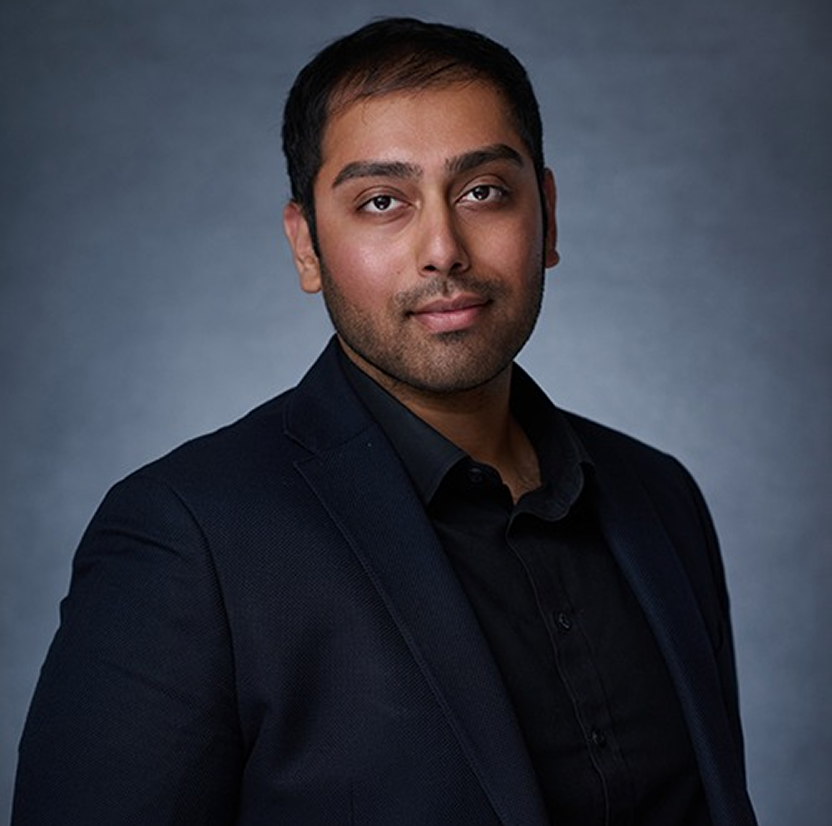 Portrait of a man with short dark hair and trimmed beard wearing a black suit jacket and black shirt, standing against a gray background.